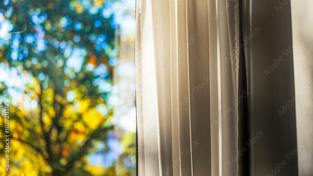 Close-up of curtains and drapes in the interior. A beautiful view of the blurred greenery outside the window in the autumn season. The light was breaking into the interior. Autumn colors