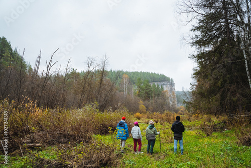 A diverse group of people is leisurely walking together through a beautiful grassy field that is surrounded by lush green woods and trees, enjoying the natural landscape around them