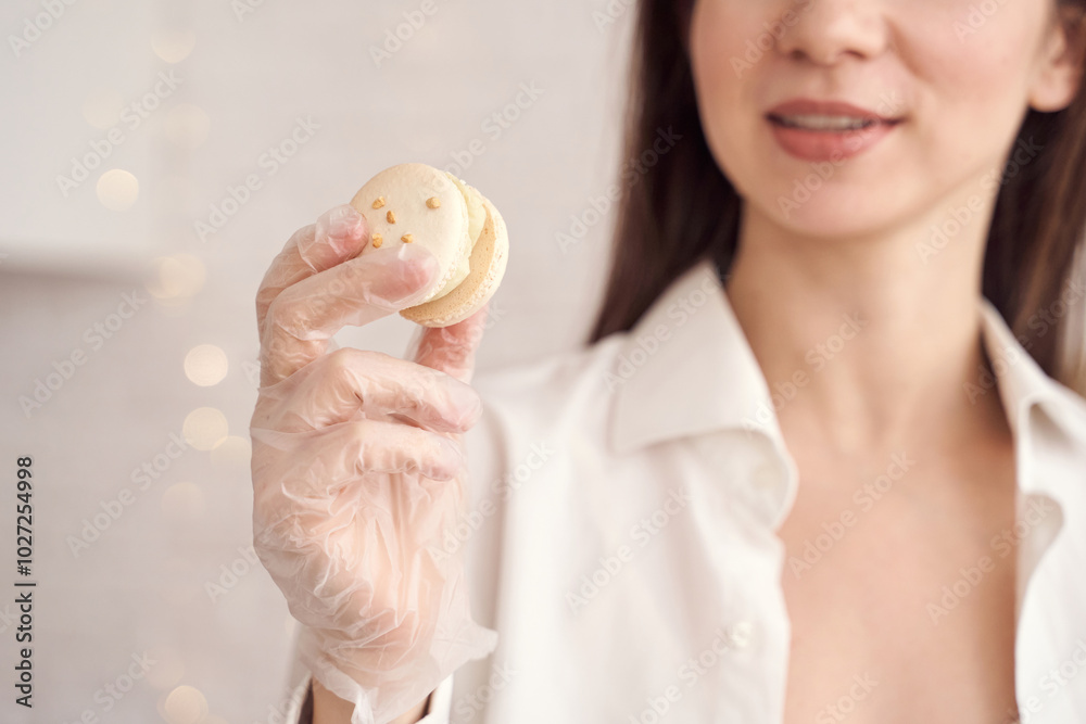 Smiling woman in white shirt holding delicious macaron with gloved hand in soft light kitchen setting. Smiling woman in white shirt holding delicious macaron with gloved hand in soft light kitchen setting.