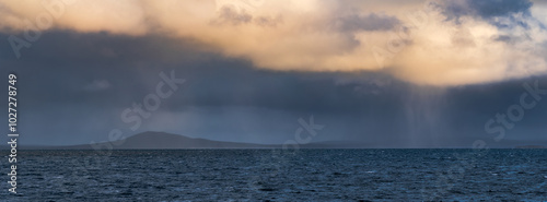 Panoramic view of rain falling from clouds in a dark sky over a distant coastline