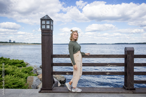 young blond woman in a sweater and trousers stands on a bridge on the lake, smiles, poses, photosession photo