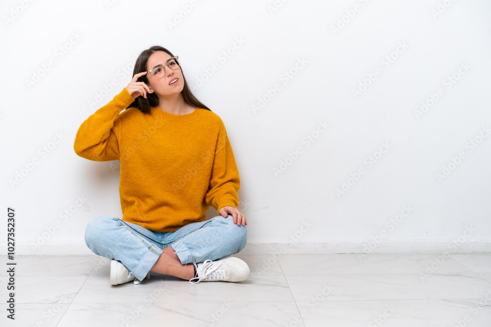Young girl sitting on the floor isolated on white background having doubts and with confuse face expression Young girl sitting on the floor isolated on white background having doubts and with confuse face expression