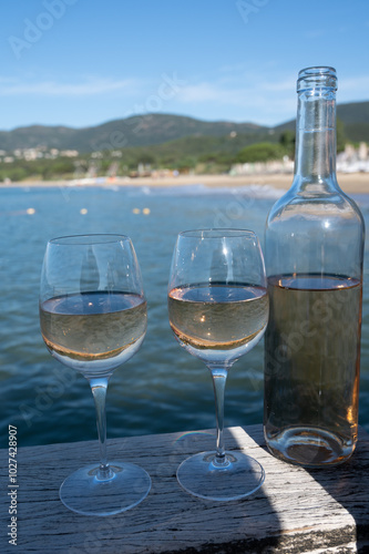 Bottle and glass of cold rose wine from Provence and wooden yacht boota pier on white sandy beach Plage de Pampelonne near Saint-Tropez, summer vacation in France photo