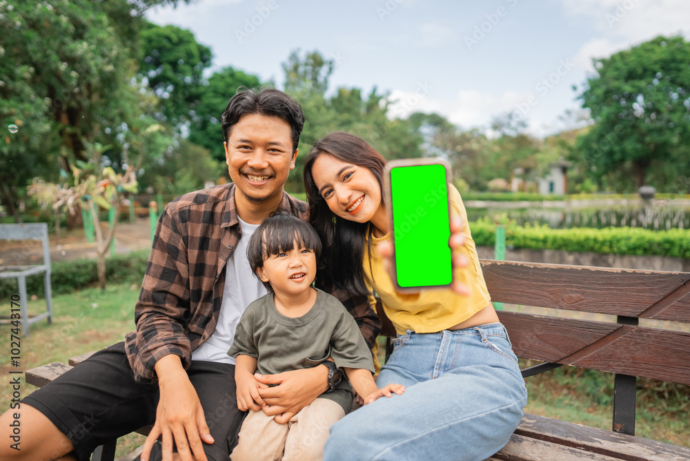 young mother shows phone screen while sitting with husband and little boy on park bench young mother shows phone screen while sitting with husband and little boy on park bench