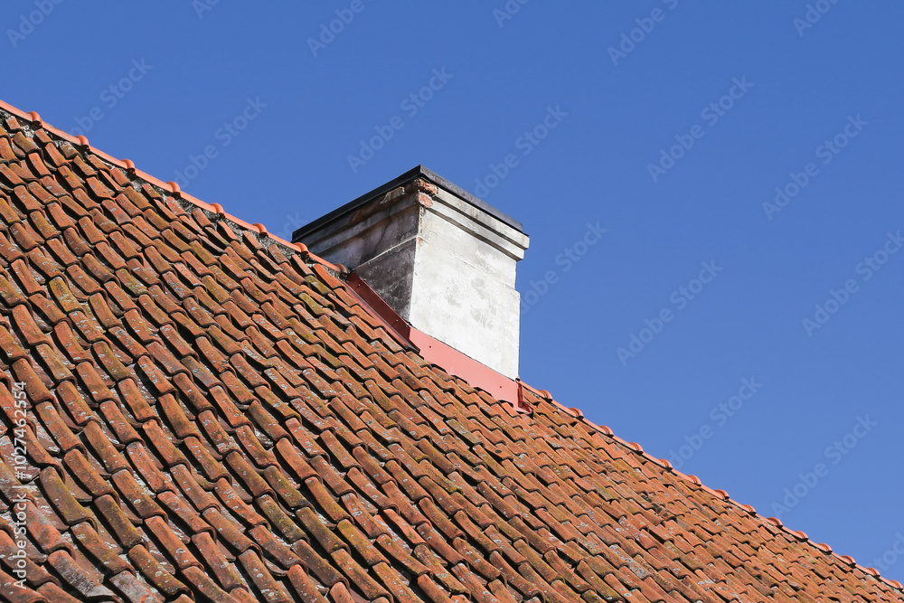 Old orange ceramic roof tiles. Tiled roof texture. Blue sky over the house. Chimney wooden vent structure at the rooftop's peak. Rustic roof covered in mold. Uneven roofing construction. Old orange ceramic roof tiles. Tiled roof texture. Blue sky over the house. Chimney wooden vent structure at the rooftop's peak. Rustic roof covered in mold. Uneven roofing construction.