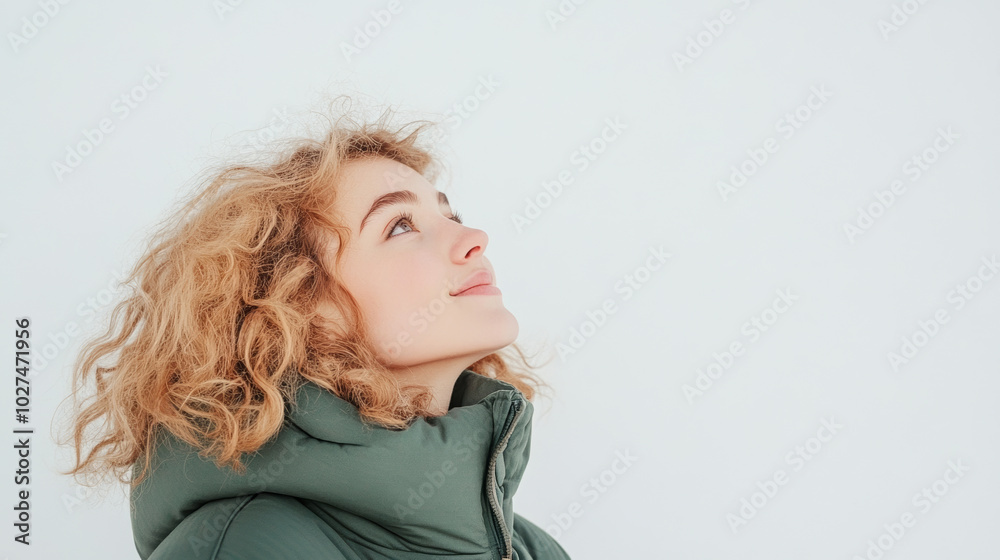 A young woman with curly hair gazes upward, wearing green puffer jacket against light background. Her expression reflects curiosity and wonder, creating serene atmosphere A young woman with curly hair gazes upward, wearing green puffer jacket against light background. Her expression reflects curiosity and wonder, creating serene atmosphere