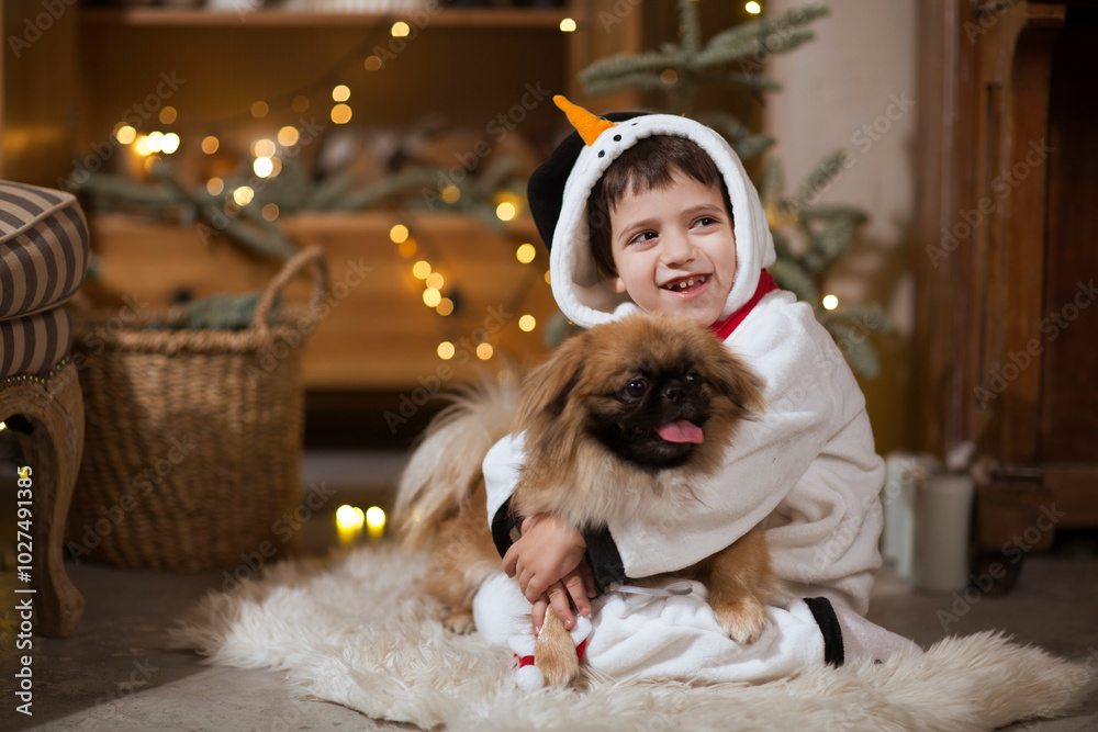 Obraz premium Happy childin snowman pyjamas embraces dog Pekingese celebrates Christmas at home. Christmas time.