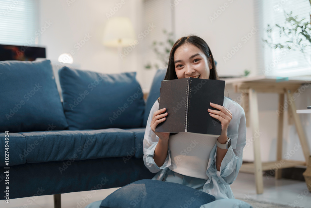 Young woman is sitting on the floor in her living room, holding a notebook in front of her face and smiling Young woman is sitting on the floor in her living room, holding a notebook in front of her face and smiling