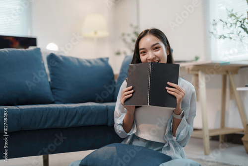 Young woman is sitting on the floor in her living room, holding a notebook in front of her face and smiling