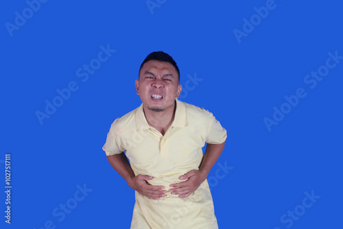 Funny face of asian man wearing t-shirt suffering from stomachache due to diarrhea on blue background

