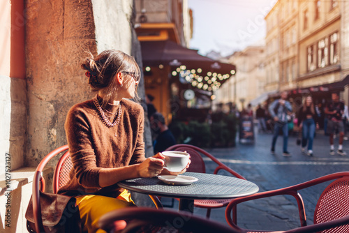 Portrait of stylish woman sitting in cafe outdoor drinking coffee. Woman enjoying architecture of Lviv city on street