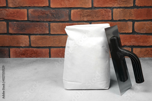 Bag of cement and plastering trowel on light textured table against red brick wall. Space for text