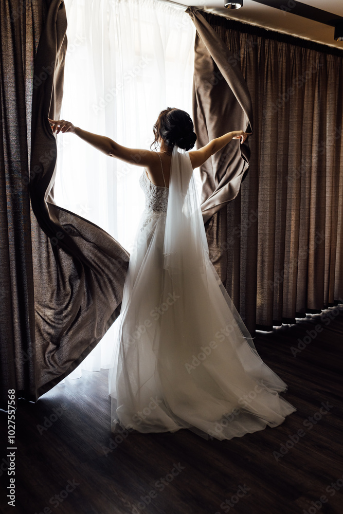 Fototapeta premium The girl stands with her back near the window and opens the curtains with her hands
