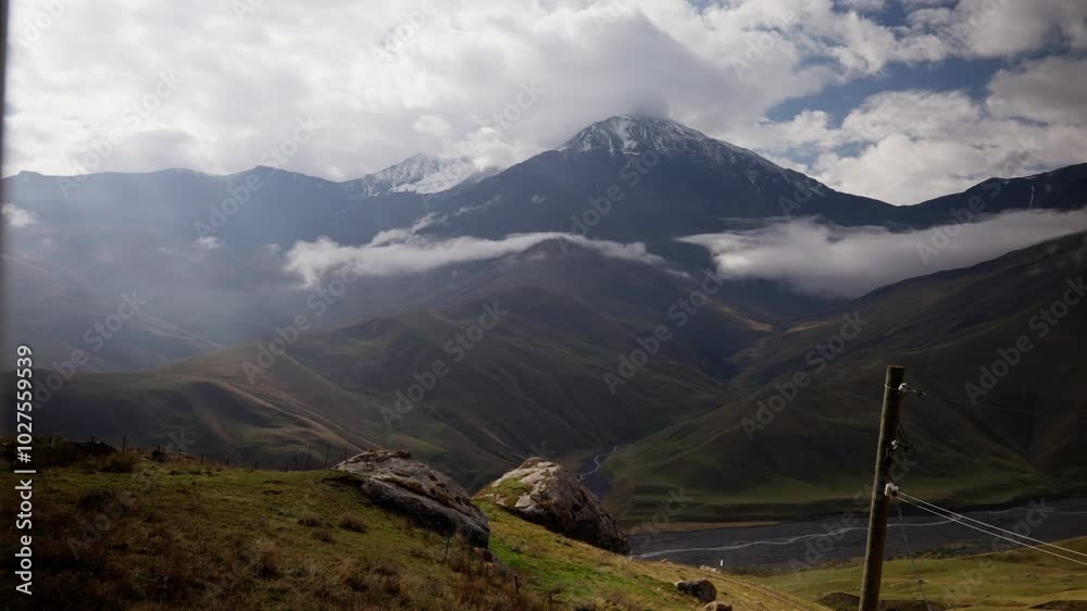 Sunlight illuminates a green valley with a lake as clouds move, revealing a snow-capped mountain peak in the background, creating a stunning natural landscape Sunlight illuminates a green valley with a lake as clouds move, revealing a snow-capped mountain peak in the background, creating a stunning natural landscape