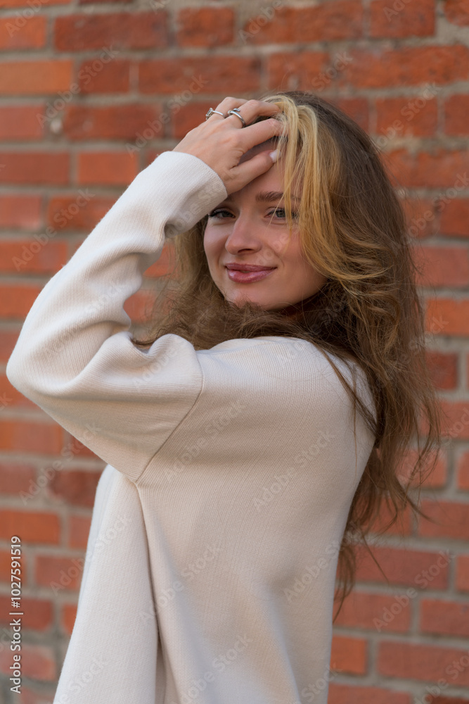 Close-up portrait of beautiful long haired caucasian blond woman in white sweater standing outdoors by red brick loft building wall. Woman looking at camera. Fashion and beautiful people theme.