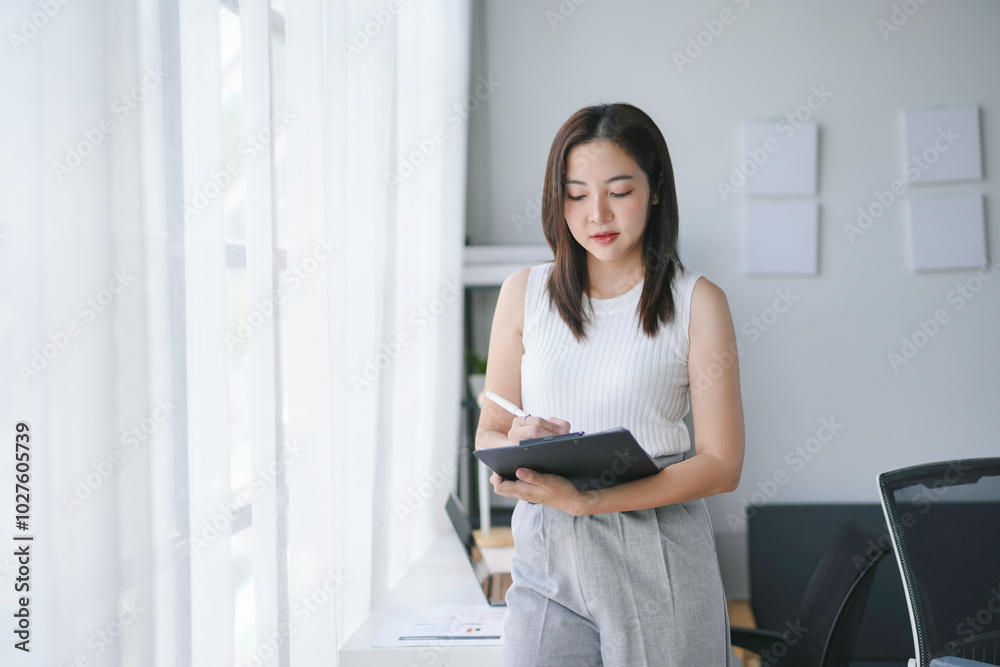 Businesswoman is standing near the window and taking notes on a clipboard during her workday in the office Businesswoman is standing near the window and taking notes on a clipboard during her workday in the office