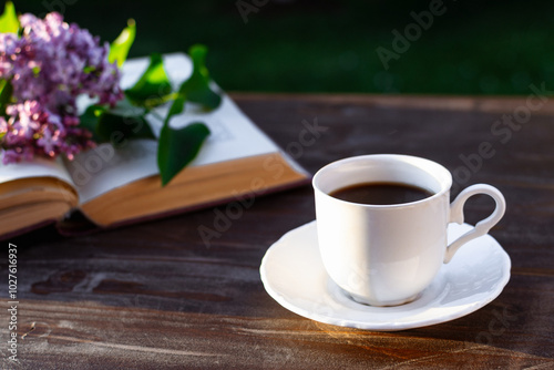 Little white cup of espresso coffee, opened bookwith purple lilac flowers on rustic wooden table in the garden at spring morning.