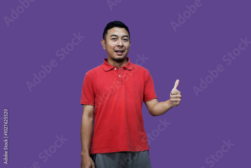Excited Asian man wears red clothes, gives thumbs-up hand gesture of approval, isolated by a purple background.
