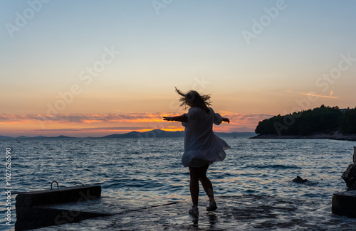 Konstfotografi Carefree woman with her hair fluttering in the wind and in a flowing dress dancing by the pier at sunset