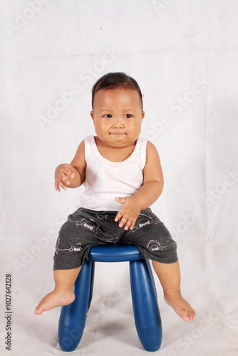 Adorable one year old Asian baby boy is sitting on a plastic chair wearing an undershirt