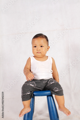 Adorable one year old Asian baby boy is sitting on a plastic chair wearing an undershirt