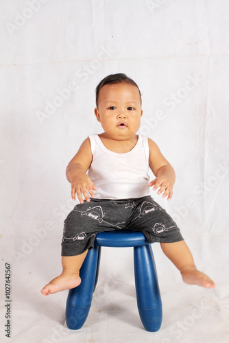 Adorable one year old Asian baby boy is sitting on a plastic chair wearing an undershirt