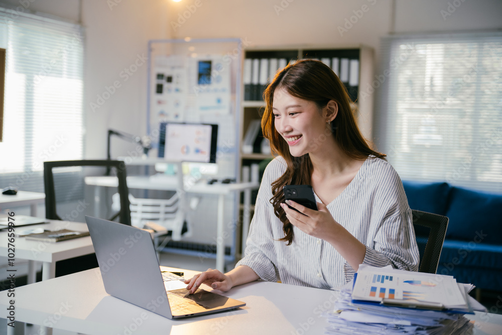 Smiling businesswoman is working on a laptop and using a smartphone at her desk in a modern office. She is surrounded by paperwork and digital devices Smiling businesswoman is working on a laptop and using a smartphone at her desk in a modern office. She is surrounded by paperwork and digital devices