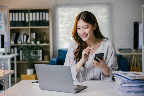 Smiling businesswoman using laptop and smartphone in a modern office, showcasing joy and positivity while managing tasks efficiently