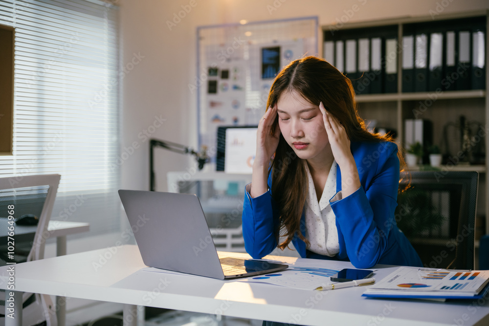 Young businesswoman is having a headache and feeling tired from working on a laptop computer at the office. She is massaging her head to relieve stress Young businesswoman is having a headache and feeling tired from working on a laptop computer at the office. She is massaging her head to relieve stress