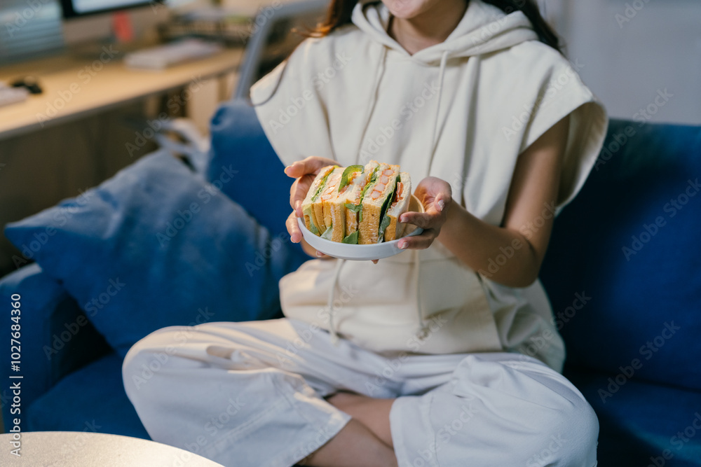 Young woman wearing casual clothes is sitting on a sofa in her living room and holding a plate with a delicious sandwich Young woman wearing casual clothes is sitting on a sofa in her living room and holding a plate with a delicious sandwich