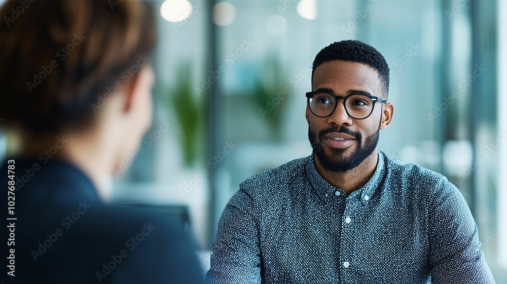 A man in glasses smiles during a conversation in a modern office setting with glass walls. A man in glasses smiles during a conversation in a modern office setting with glass walls.