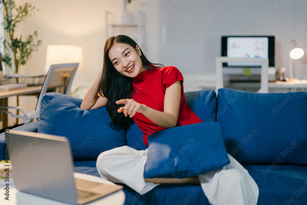 Young woman is sitting on a comfortable blue sofa in her living room, enjoying a relaxing evening using her laptop for entertainment Young woman is sitting on a comfortable blue sofa in her living room, enjoying a relaxing evening using her laptop for entertainment