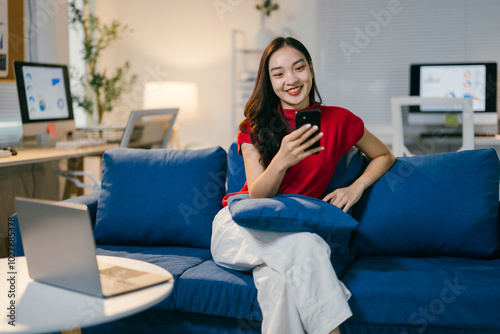 Young asian woman is sitting on a sofa in her home office, using a smartphone while smiling. She is wearing a red shirt and white pants