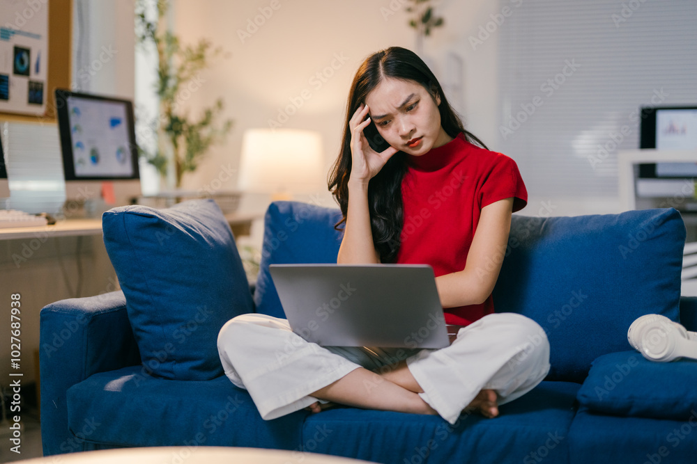 Young asian businesswoman is sitting on her couch at home, working on her laptop. She is feeling stressed and overwhelmed by her work Young asian businesswoman is sitting on her couch at home, working on her laptop. She is feeling stressed and overwhelmed by her work