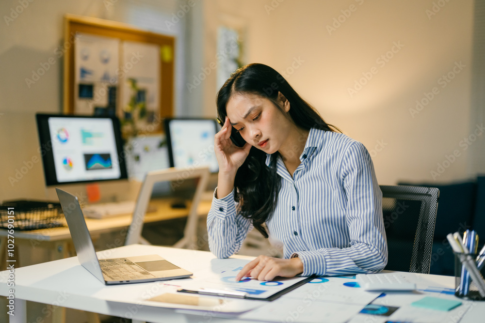 Businesswoman is feeling stressed and having a headache while working late at her desk in the office, surrounded by financial documents and multiple computer screens Businesswoman is feeling stressed and having a headache while working late at her desk in the office, surrounded by financial documents and multiple computer screens