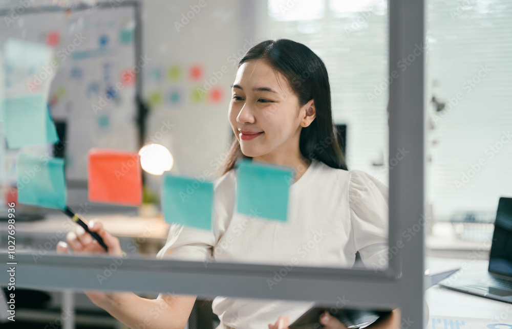 Young asian businesswoman is using a marker to write on sticky notes while brainstorming new project ideas in a modern office Young asian businesswoman is using a marker to write on sticky notes while brainstorming new project ideas in a modern office