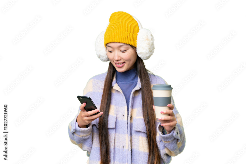 Young Asian woman wearing winter muffs over isolated chroma key background holding coffee to take away and a mobile Young Asian woman wearing winter muffs over isolated chroma key background holding coffee to take away and a mobile