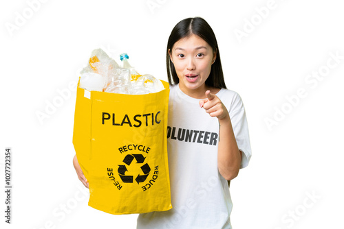 Young Asian woman holding a bag full of plastic bottles to recycle over isolated chroma key background surprised and pointing front