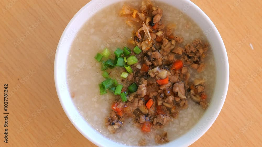 Close-up view of delicious bowl of rice porridge garnished with minced meat, vegetables, and green onions on wooden table, showcasing traditional Asian cuisine. Traditional Asian Food. Close-up view of delicious bowl of rice porridge garnished with minced meat, vegetables, and green onions on wooden table, showcasing traditional Asian cuisine. Traditional Asian Food.