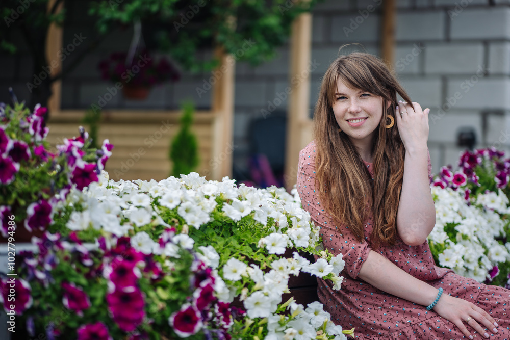 Young woman with flowing brown hair smiles as she adjusts her hair while sitting among white and purple flowers on a bench Young woman with flowing brown hair smiles as she adjusts her hair while sitting among white and purple flowers on a bench