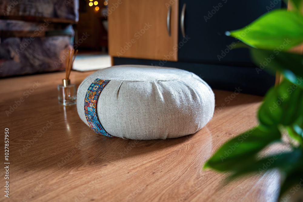 Side angle view of a grey meditation zafu cushion with a colorful stripe sits on a wooden floor, next to a perfume sticks inside a house against couch and lights, cozy interior of a house