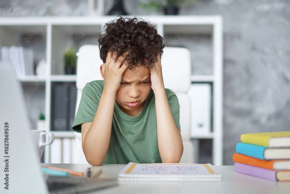 Cute little sad African American schoolboy sitting at the table holding his head in despair, tired of learning his home assignment