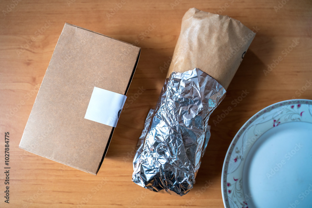 Top view of a foil-wrapped burrito next to a cardboard takeout box on a wooden table, capturing the essence of a convenient meal or takeout experience.