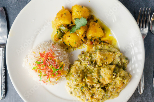 Deshi plate of fluffy colorful rice topped with bits of red and yellow, with golden boiled potatoes seasoned with herbs, greenish chciken curry with vegetables. photo