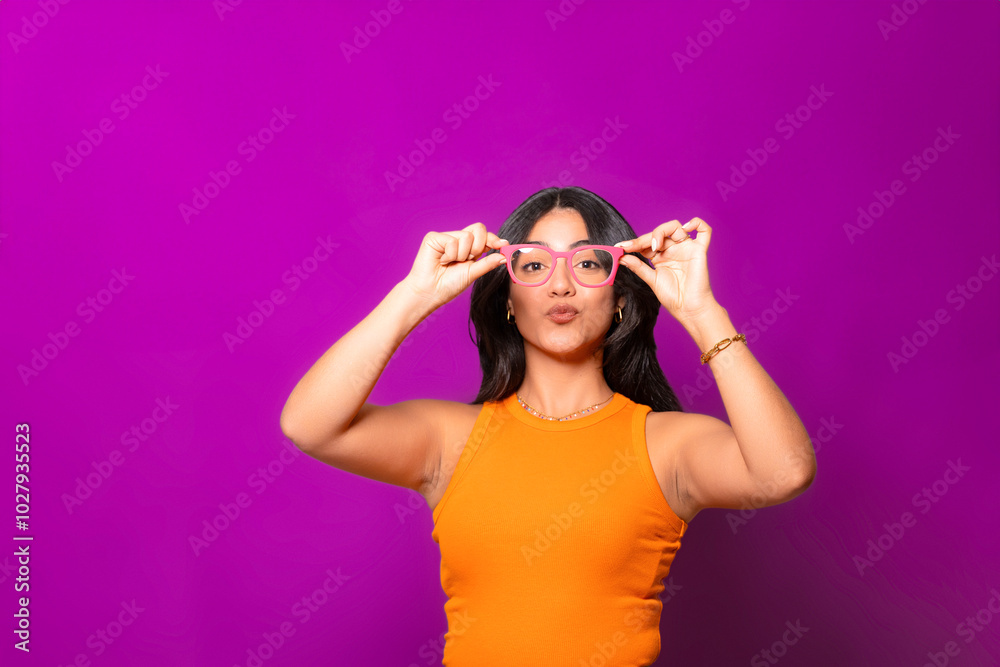 Playful woman making a duck face, holding pink glasses in both hands, wearing an orange top on a vibrant purple background. Playful woman making a duck face, holding pink glasses in both hands, wearing an orange top on a vibrant purple background.