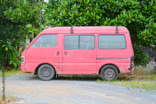 A rusty red van is parked on a dirt road, surrounded by lush greenery.