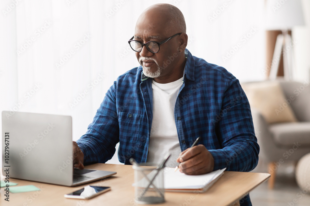 Senior Black Man Using Laptop And Writing Report Taking Notes Working Online Sitting At Desk In Office, Wearing Eyeglasses. Entrepreneurship In Mature Age, Business Lifestyle Concept Senior Black Man Using Laptop And Writing Report Taking Notes Working Online Sitting At Desk In Office, Wearing Eyeglasses. Entrepreneurship In Mature Age, Business Lifestyle Concept