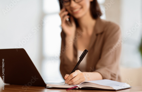 Taking Notes. Young secretary sitting at desk with laptop computer and talking on mobile phone, writing in notebook