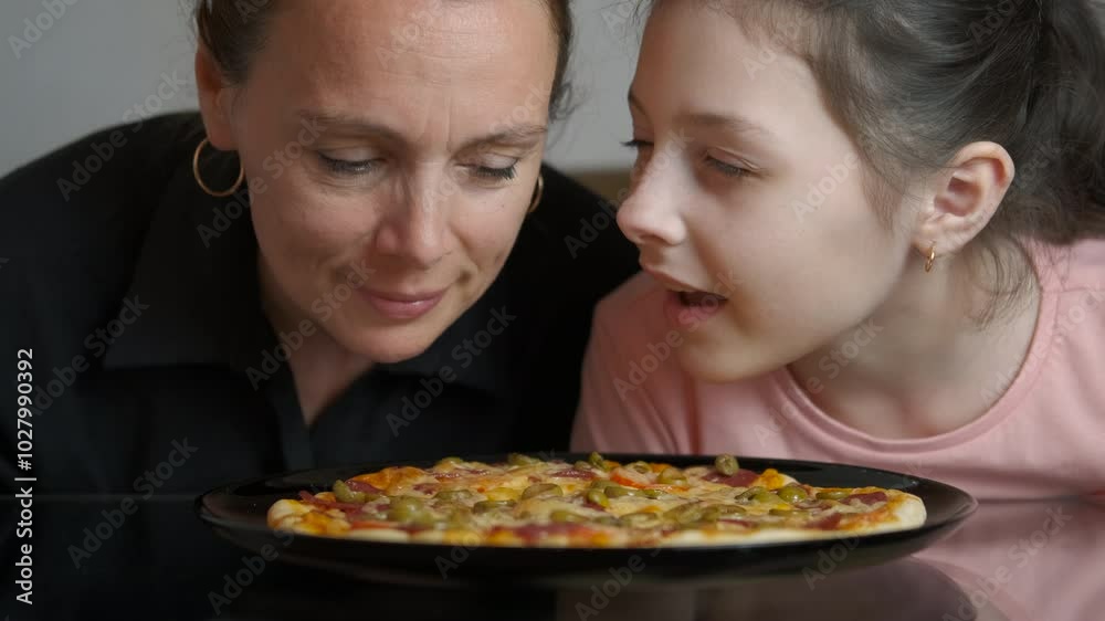 Mum and daughter look at baked pizza. A view of mum with her daughter feel the spice flavor of cooked homemade pizza o the table in the kitchen. Mum and daughter look at baked pizza. A view of mum with her daughter feel the spice flavor of cooked homemade pizza o the table in the kitchen.