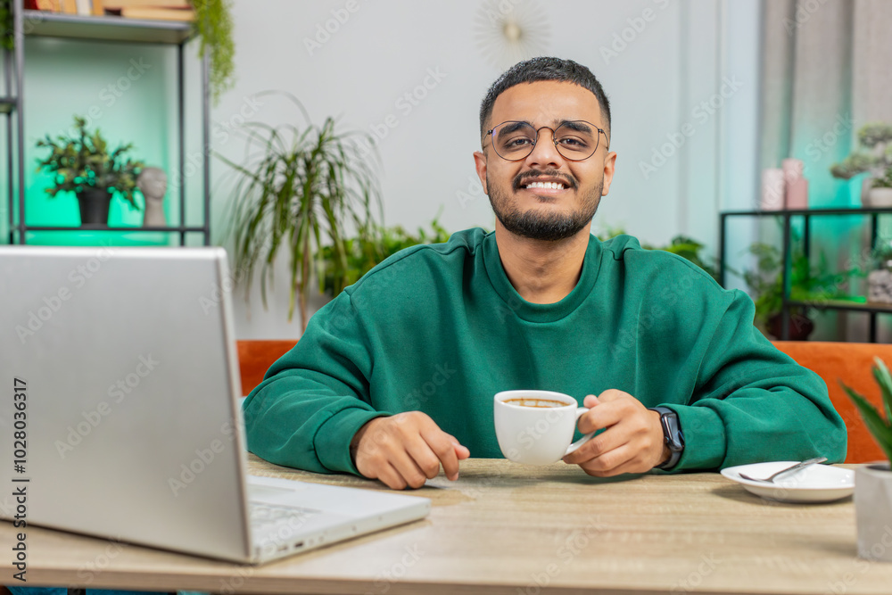 Attractive smiling Indian man working on laptop computer drinking a cup of warm hot coffee sits at table workplace home office room in the morning. Arabian Hindu guy enjoying relaxing, taking a break Attractive smiling Indian man working on laptop computer drinking a cup of warm hot coffee sits at table workplace home office room in the morning. Arabian Hindu guy enjoying relaxing, taking a break